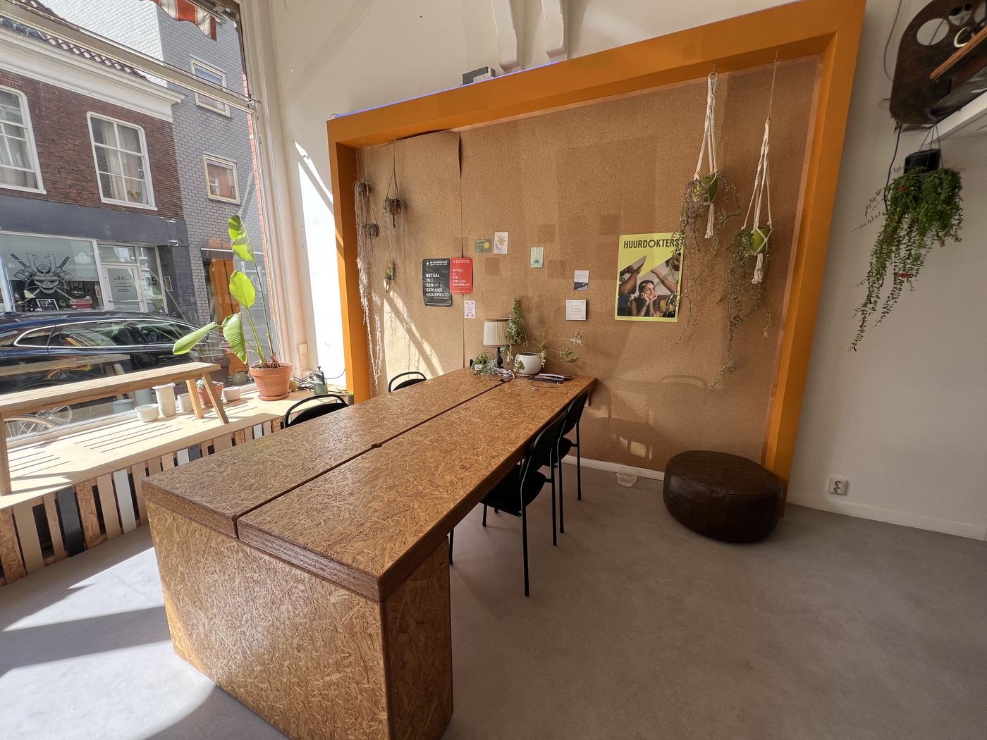 De voorruimte van Waarkstee: een grote houten eettafel voor een oranje prikbord met hangplanten, met zicht op de Nieuweweg.