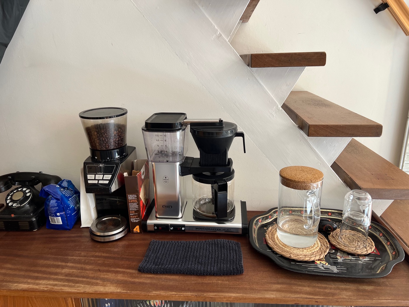 De koffiehoek onder de trap: een koffiemolen, filtermachine en glazen op een dienblad.